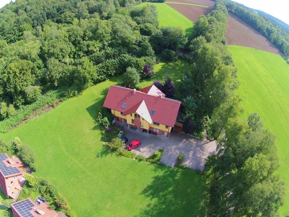 Luftaufnahme eines gelben Hauses mit rotem Dach, umgeben von grünen Rasenflächen, Bäumen und Feldern an einem sonnigen Tag.