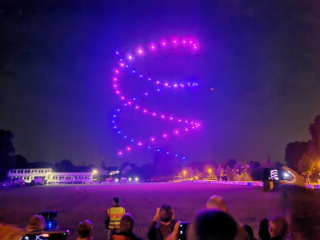 Eine Drohnen-Lichtshow erzeugt eine wirbelnde, violette Spiralform am Nachthimmel über einer Menschenmenge, die von einem Feld aus zuschaut.