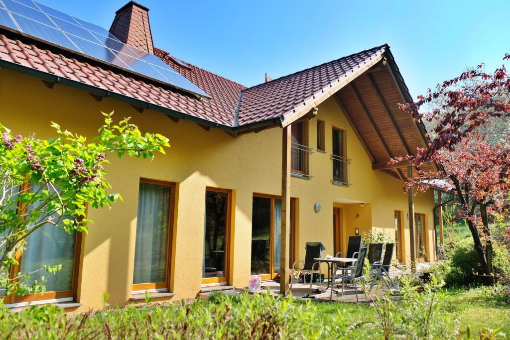 Ein zweistöckiges gelbes Haus mit Ziegeldach und Solaranlage, mit Terrasse mit Gartenmöbeln und Garten.