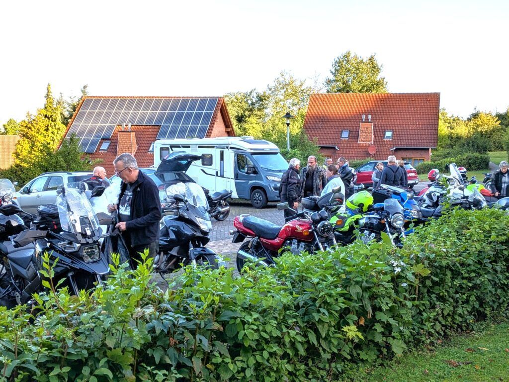 Eine Gruppe von Menschen steht neben geparkten Motorrädern und einem Wohnmobil in einem sonnigen, grünen Außenbereich mit zwei Häusern.