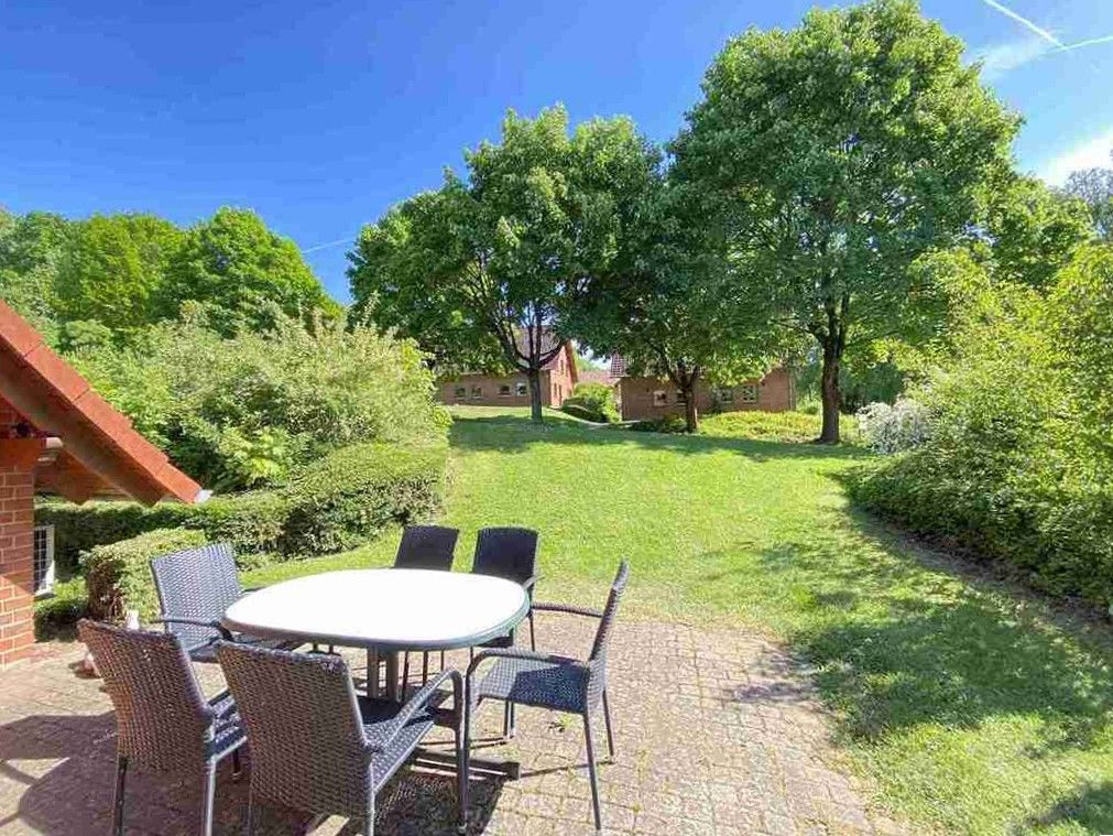 Eine Terrasse mit einem weißen runden Tisch und Stühlen, die auf einen grasbewachsenen Hof mit Bäumen unter einem klaren blauen Himmel blickt.