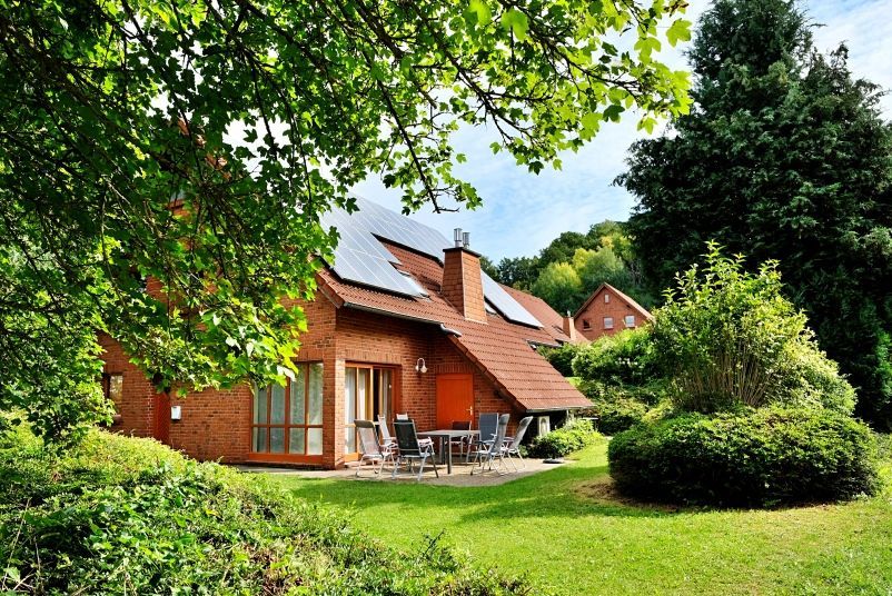 An einem sonnigen Tag steht ein Haus aus roten Backsteinen mit braunem Ziegeldach und Solarpaneelen in einem üppig grünen Garten.