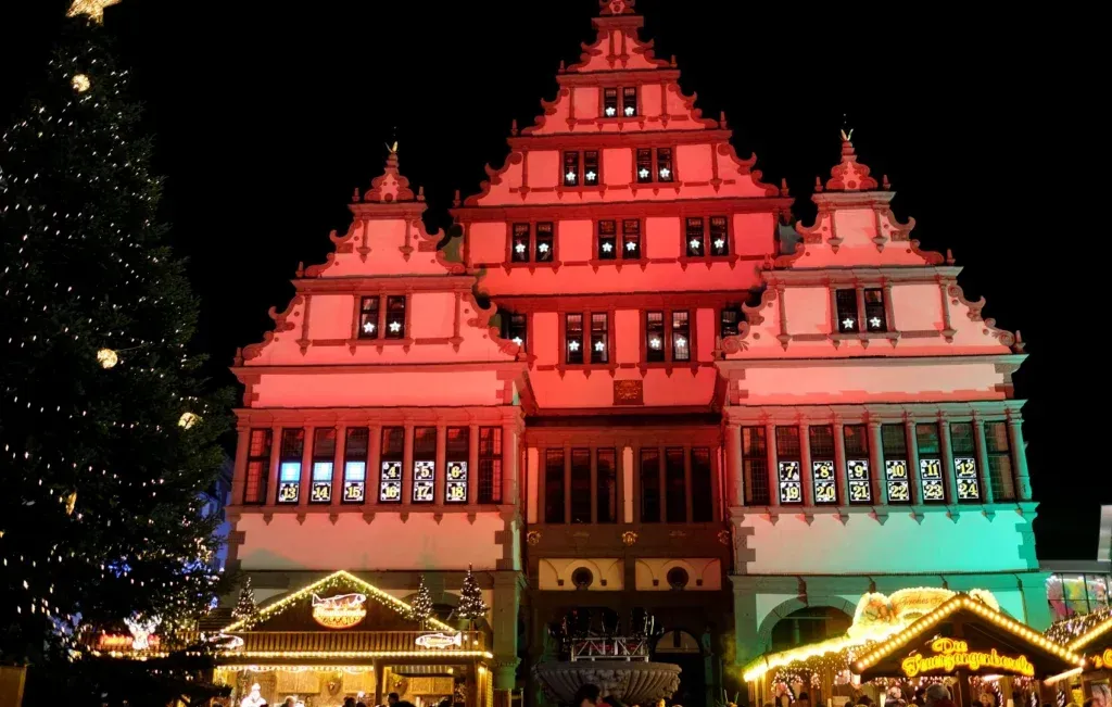 Ein beleuchtetes historisches Gebäude mit gestaffelter Giebelfassade erstrahlt in rotem Licht, flankiert von einem Weihnachtsbaum und einem Markt.