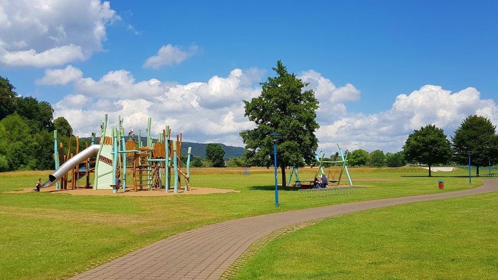 Ein Spielplatz mit Klettergerüst, Rutsche und Schaukel auf einer grünen Wiese unter blauem Himmel mit flauschigen weißen Wolken.