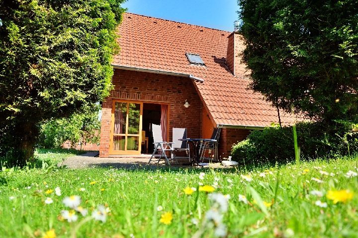 Ein Ferienhaus aus Backstein mit rotem Ziegeldach und Gartenmöbeln auf der Terrasse, eingebettet in einen üppigen Garten mit gelben Blumen.