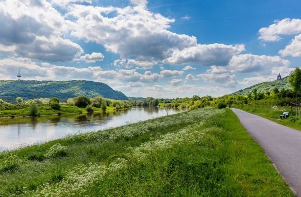 Ein gepflasterter Weg verläuft entlang eines ruhigen Flusses, gesäumt von grasbewachsenen Ufern und Wildblumen, vor dem Hintergrund grüner Hügel unter einem bewölkten Himmel.