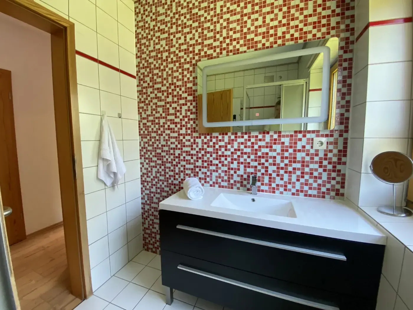 Ein Badezimmerwaschtisch mit weißer Arbeitsplatte und schwarzen Schubladen vor einer rot-weißen Mosaikfliesenwand und einem Spiegel.