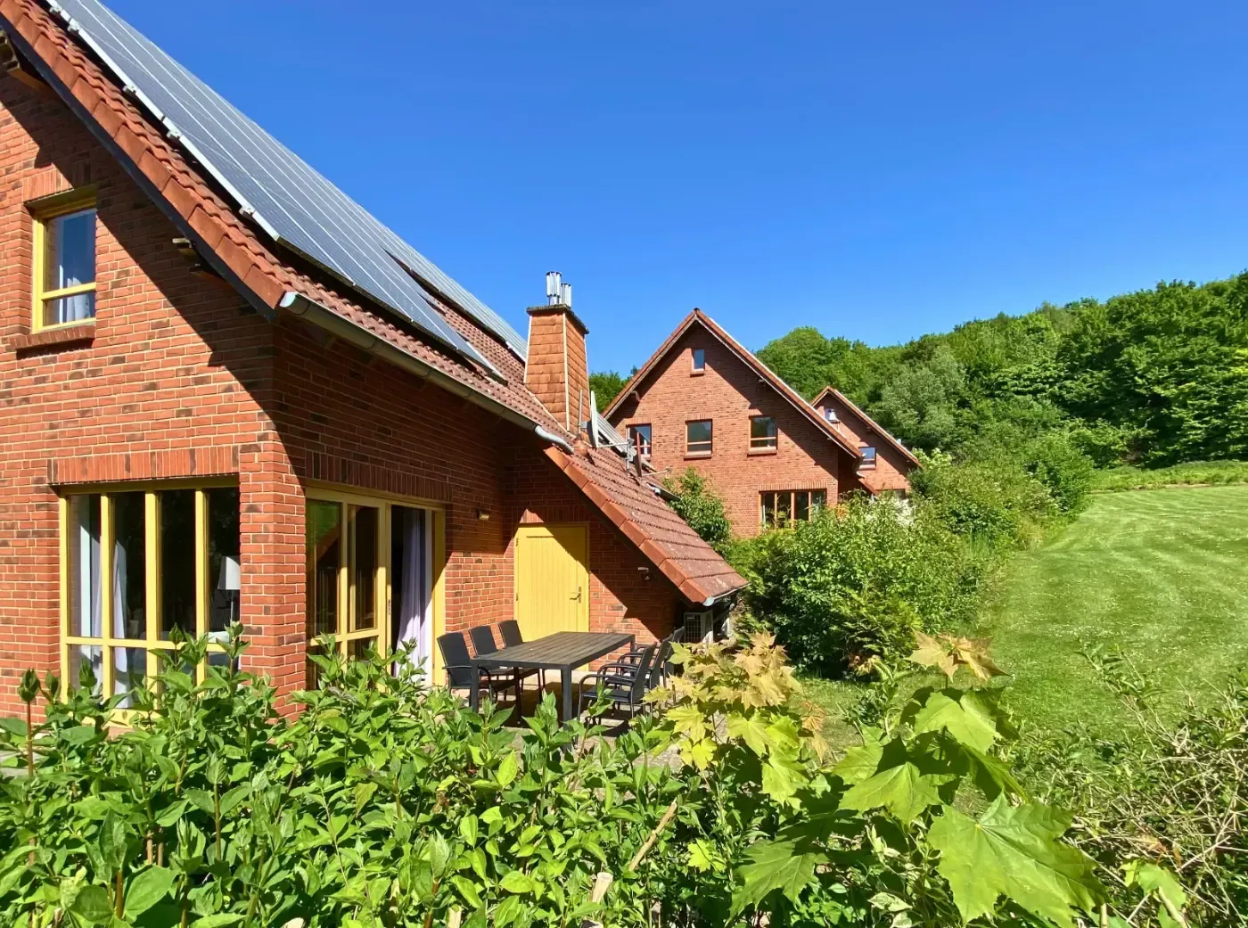 Zwei rote Backsteinhäuser mit Solarpaneelen auf den Dächern, eingebettet in üppiges Grün und einen grasbewachsenen Hang.