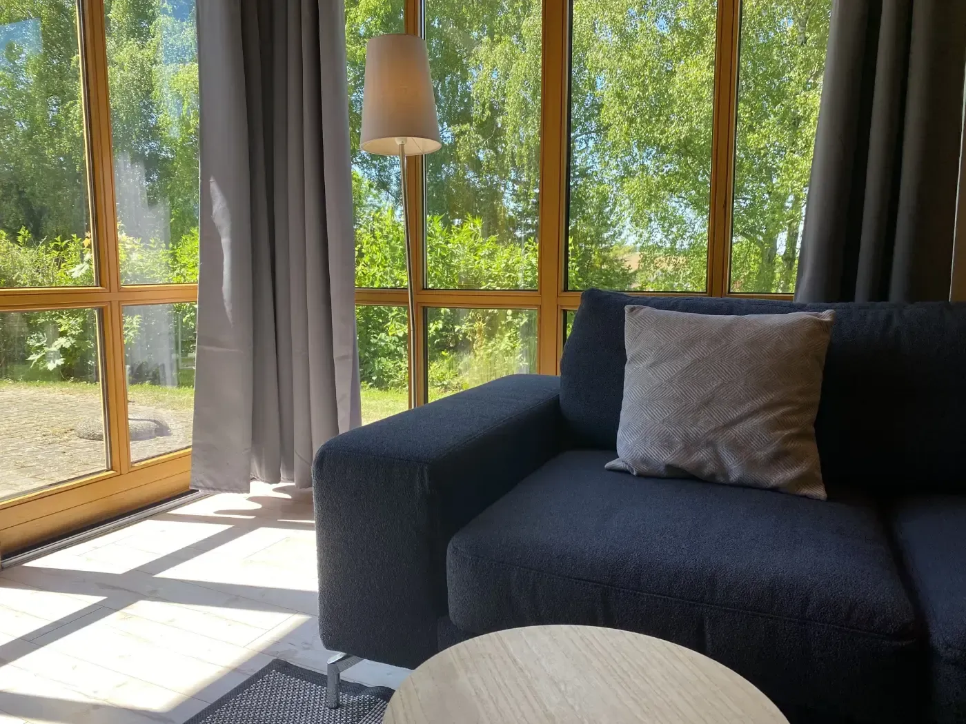 In einem sonnendurchfluteten Raum mit bodentiefen Fenstern und einer Stehlampe steht ein dunkelgraues Sofa mit einem gemusterten Zierkissen.