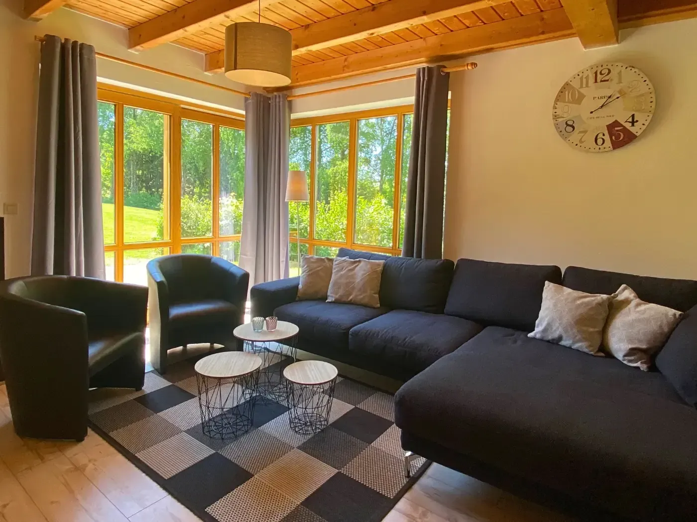 Ein gemütliches Wohnzimmer mit einem großen dunklen Sofa, zwei Sesseln, einem gemusterten Teppich und bodentiefen Fenstern mit Blick in den Garten.