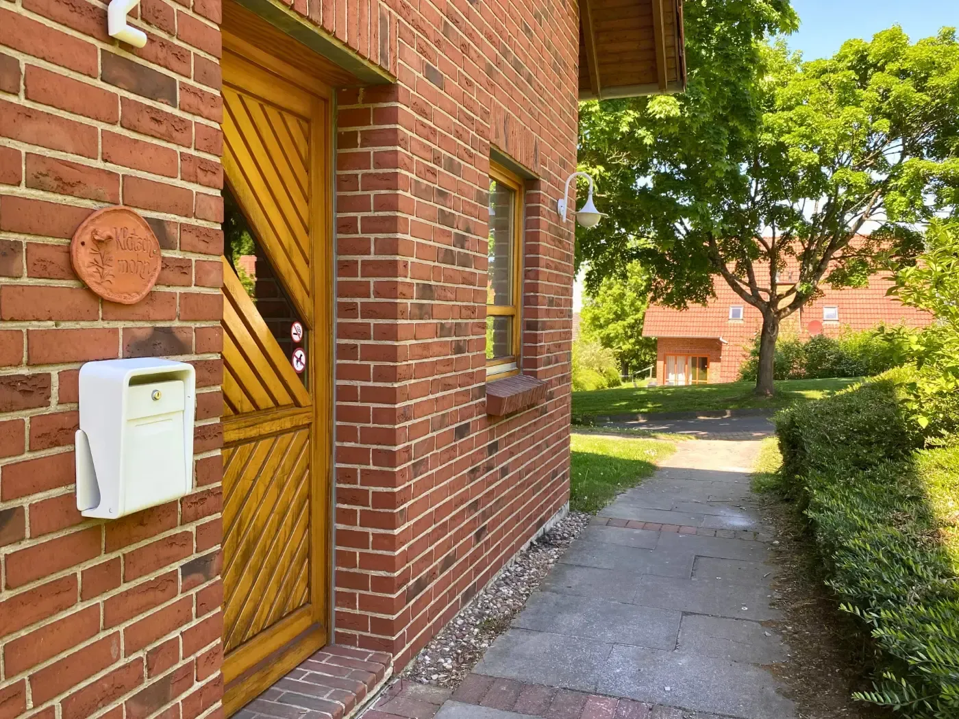 Ein gemauerter Gebäudeeingang mit einer diagonalen Holztür, einem weißen Briefkasten und einem Steinweg, der zu einem grünen Hof führt.