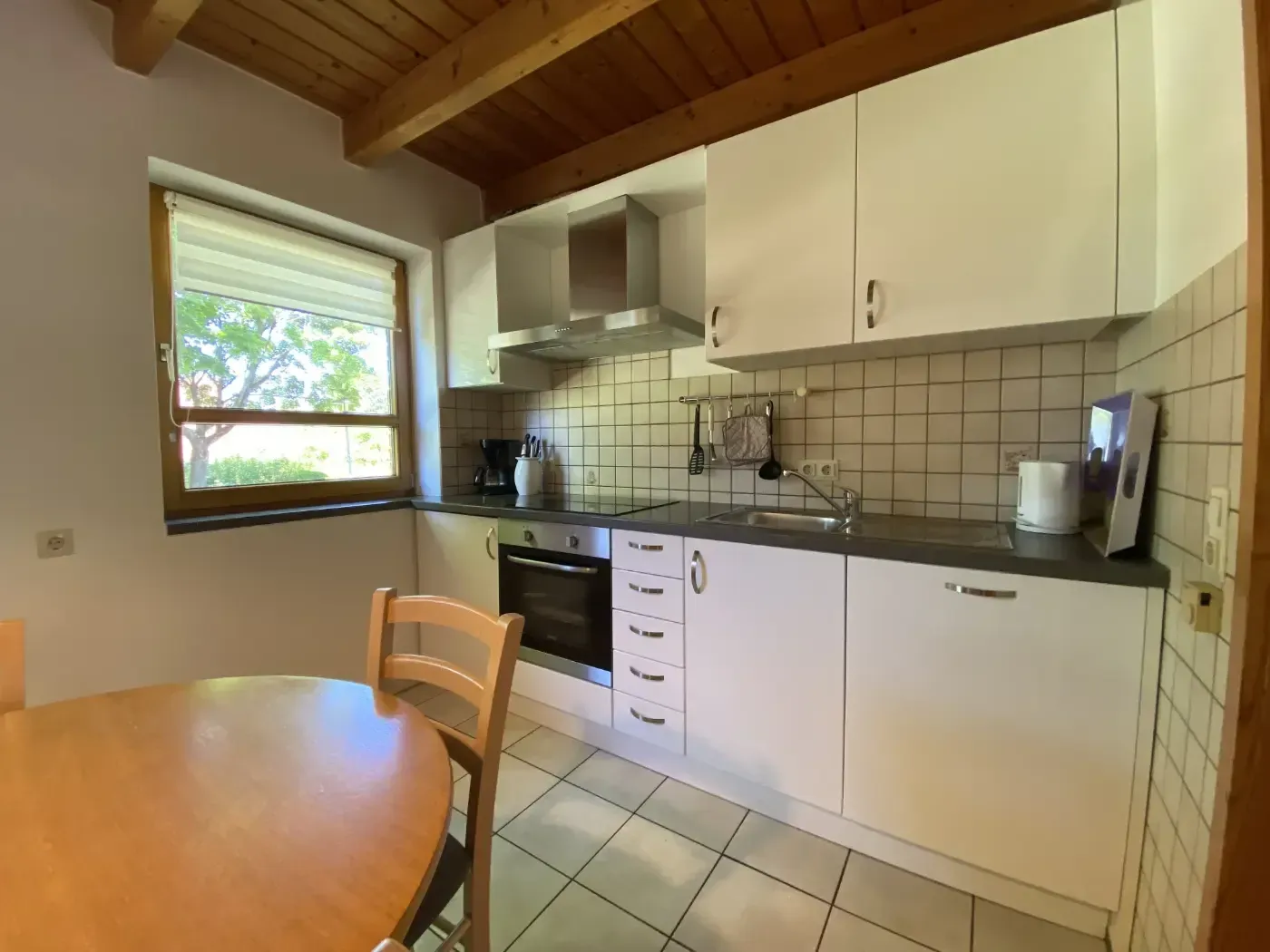 Eine kleine, helle Küche mit weißen Schränken, hellen Holzbalken an der Decke, gefliesten Wänden und einem runden Esstisch aus Holz.