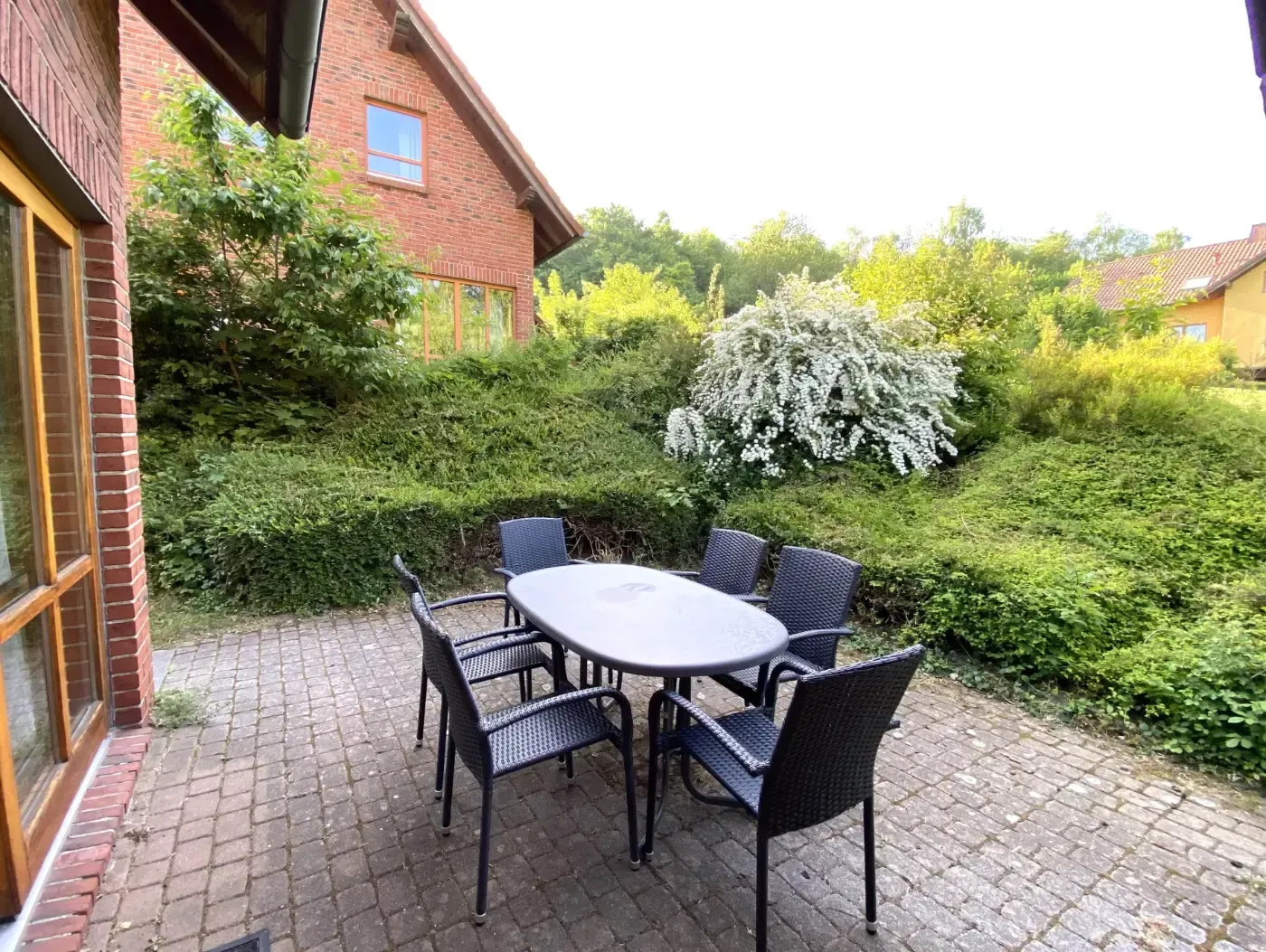 Auf einer gepflasterten Terrasse neben einem Backsteinhaus und üppiger grüner Landschaft steht eine Terrassengarnitur mit einem ovalen Tisch und vier Stühlen.