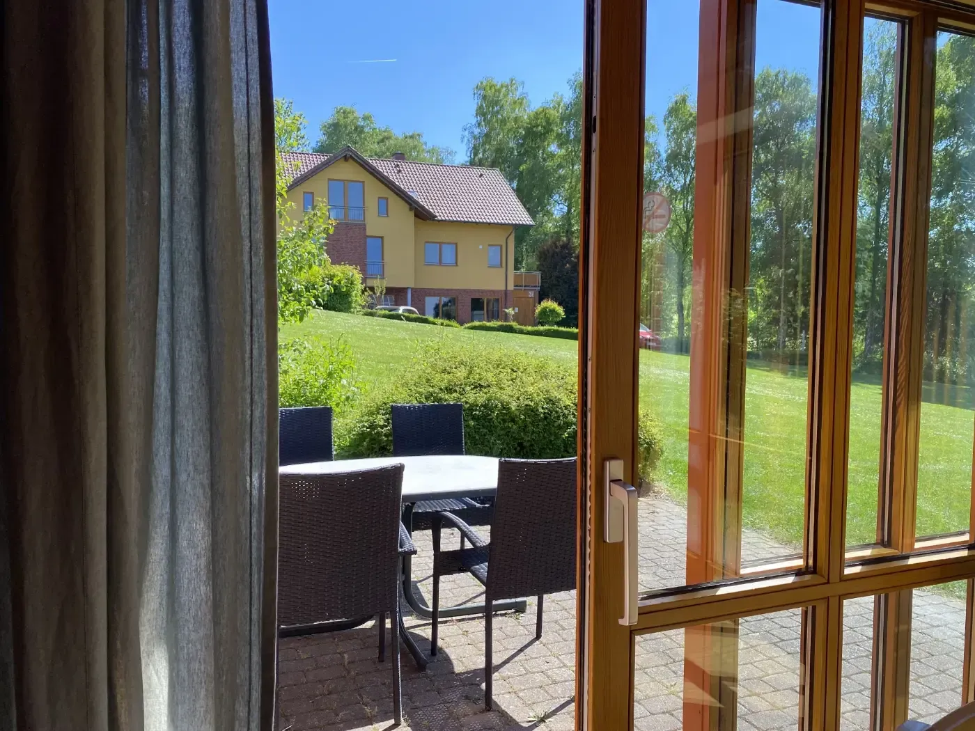 Eine Terrasse mit dunklen Korbstühlen und einem Tisch, durch eine Glastür zu sehen, mit Blick auf einen Rasen und ein gelbes Haus.