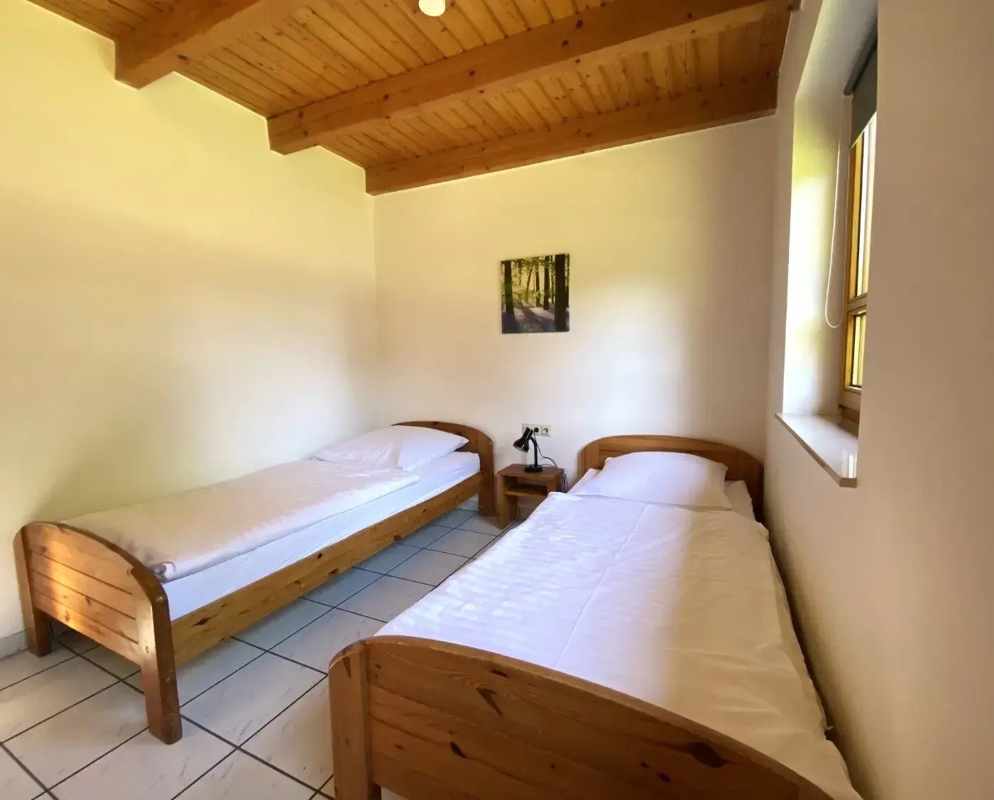 Ein Schlafzimmer mit zwei hellen Einzelbetten aus Holz, weißer Bettwäsche, hellen Wänden, Fliesenboden und einer Holzbalkendecke.