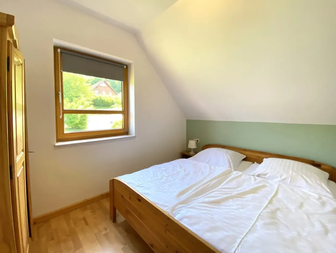 Ein Schlafzimmer mit einem hölzernen Bettgestell, weißer Bettwäsche, einem Fenster mit grauem Rollo und hellgrünen Wänden.