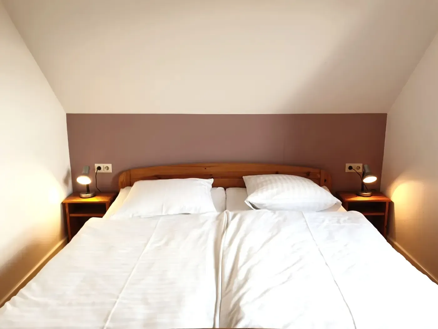 Ein ordentlich gemachtes Bett mit weißer Bettwäsche, hölzernem Kopfteil und zwei Nachttischen mit kleinen Lampen vor einer malvenfarbenen Wand.