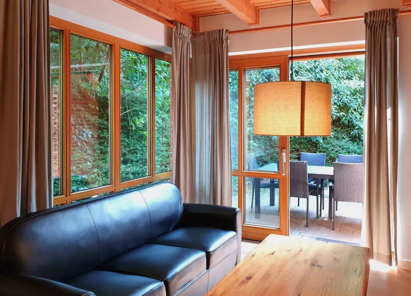 Ein schwarzes Ledersofa und ein hölzerner Couchtisch in einem sonnendurchfluteten Wohnzimmer mit Blick auf eine Gartenterrasse.