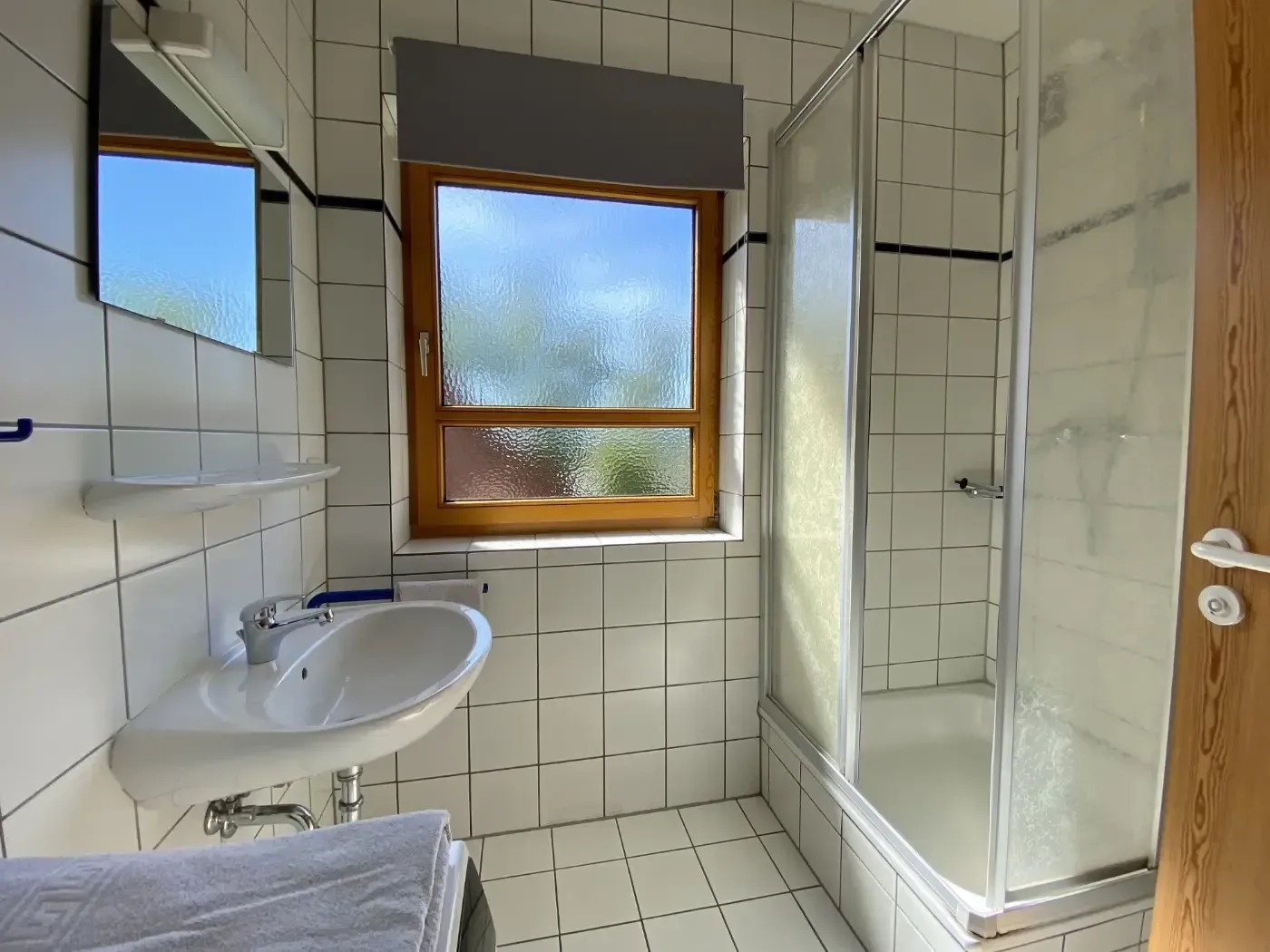 Ein gefliestes Badezimmer mit Waschbecken, verglaster Dusche, Spiegel und Fenster mit Blick ins Grüne.