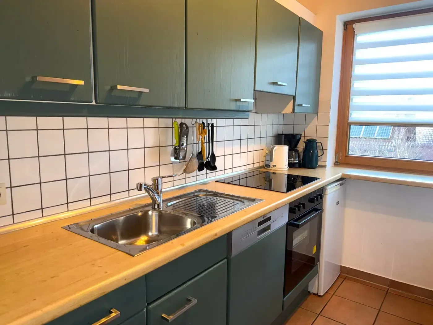 Eine Küche mit dunkelgrünen Schränken, einer holzfarbenen Arbeitsplatte, einem Edelstahlspülbecken und einem Fenster mit Jalousien.