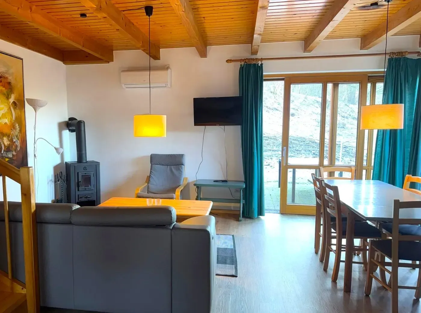 Ein gemütliches Wohnzimmer mit grauem Sofa, Holzofen, Esstisch und türkisfarbenen Vorhängen unter einer Holzbalkendecke.