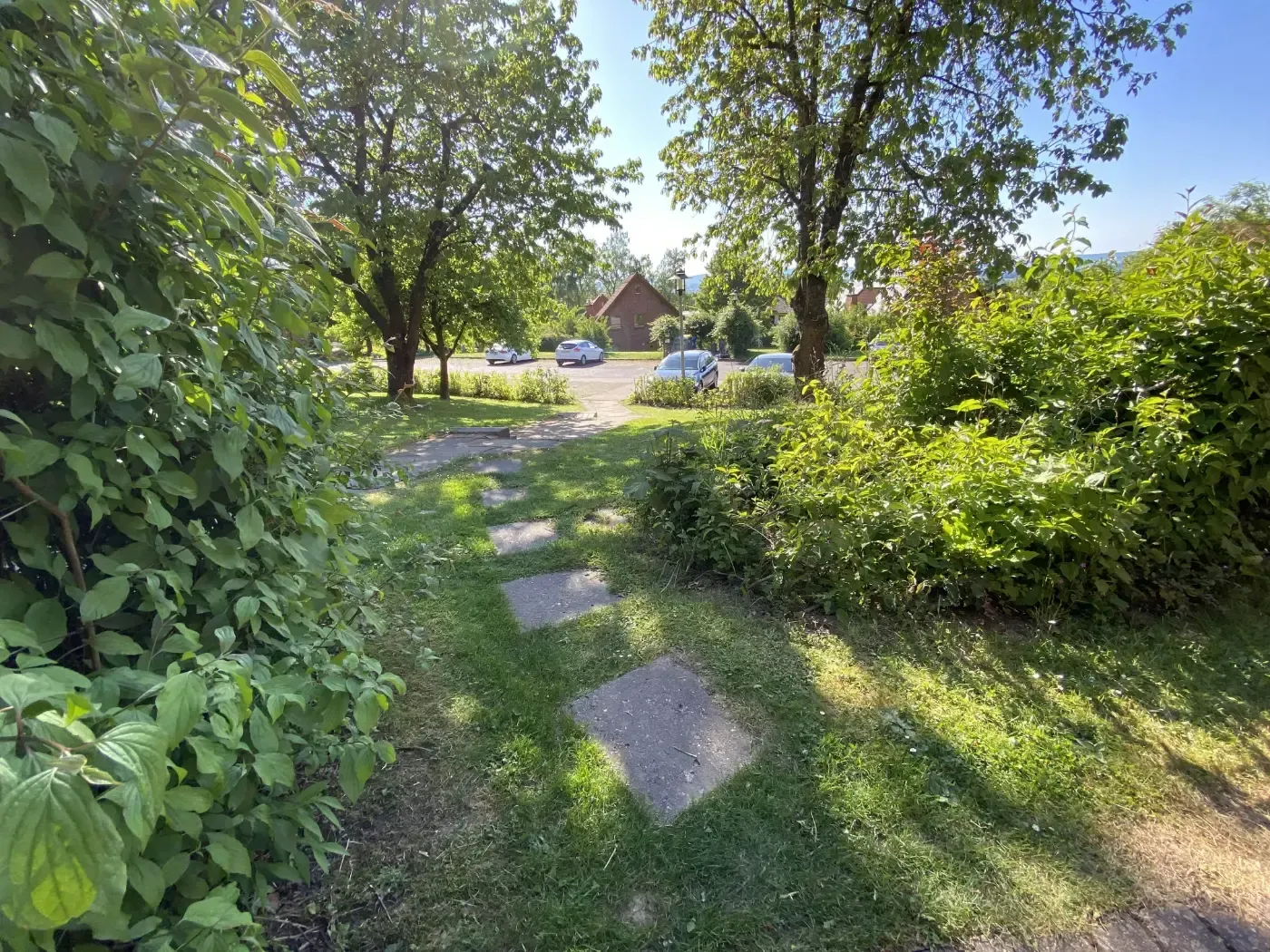 Ein gepflasterter Steinweg schlängelt sich durch einen grasbewachsenen Hof zu einem Parkplatz und Bäumen unter einem klaren blauen Himmel.