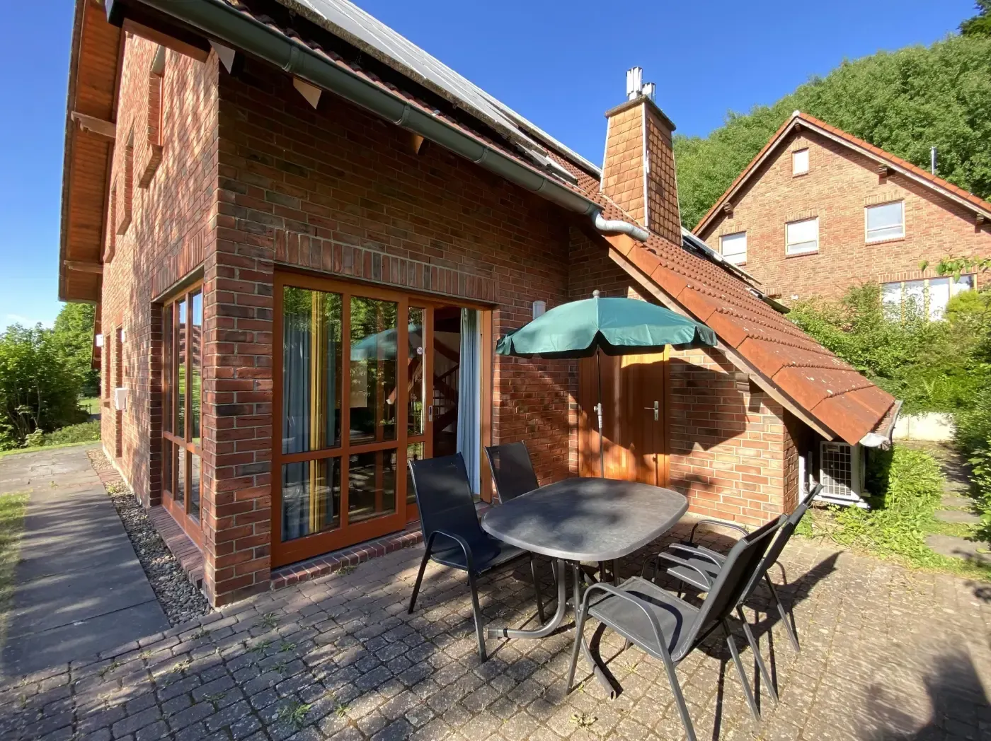 Eine sonnige Terrasse an einem Backsteinhaus mit einem kleinen Tisch, Stühlen und einem aufgespannten Sonnenschirm, vor der Kulisse grüner Bäume.