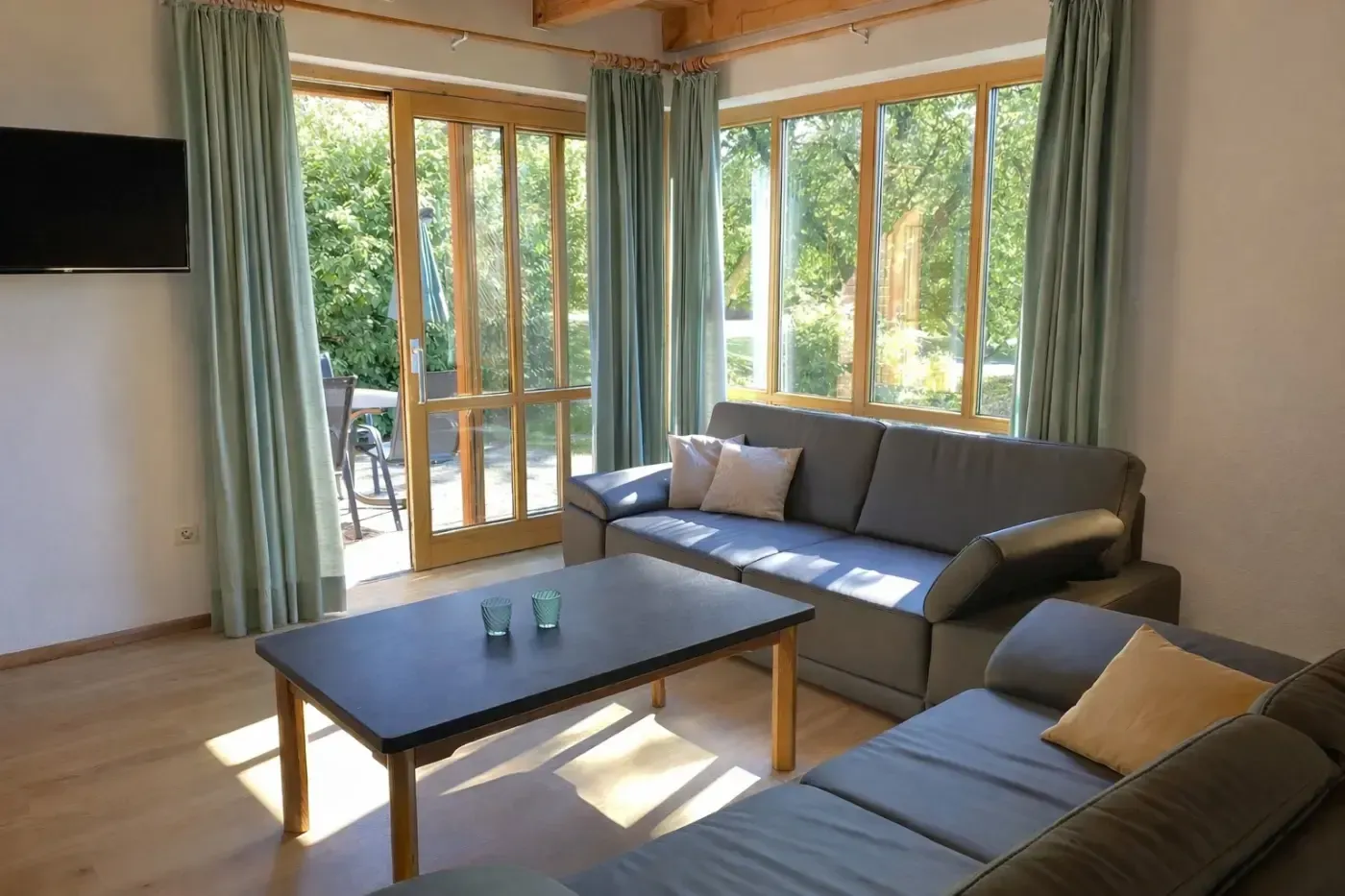 Ein helles Wohnzimmer mit zwei grauen Sofas, einem dunklen Couchtisch, hellen Holzböden und großen Fenstern mit salbeigrünen Vorhängen.