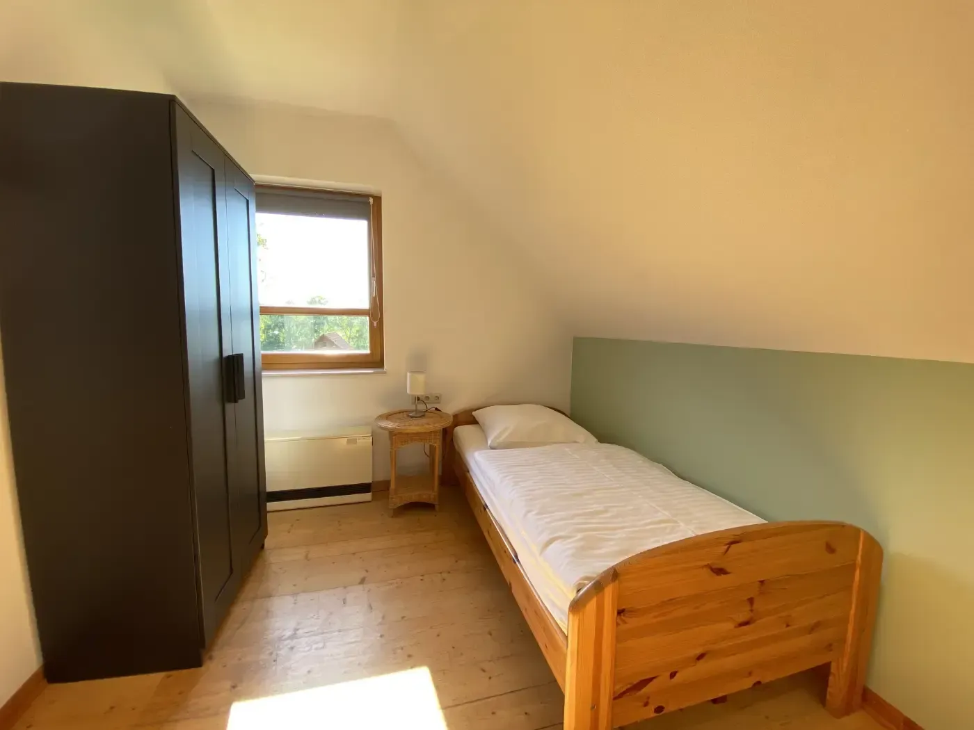 Ein Schlafzimmer mit einem hölzernen Doppelbett, einem großen schwarzen Kleiderschrank, einem kleinen Beistelltisch und einem Fenster mit Aussicht.