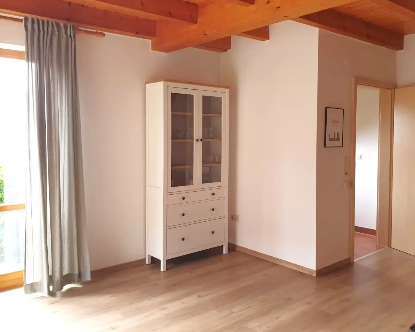 Ein weißer hölzerner Aufsatzschrank steht auf einem hellen Holzboden in einem Raum mit weißen Wänden und sichtbaren Deckenbalken.