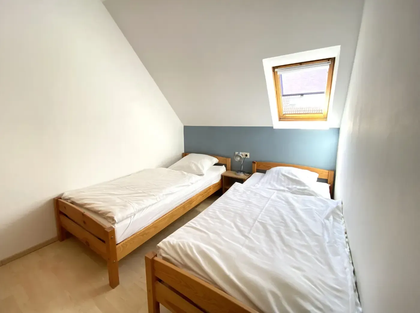 Ein Schlafzimmer mit schrägen Decken, zwei Einzelbetten aus Holz mit weißer Bettwäsche und einem kleinen Dachfenster über einer blauen Wand.