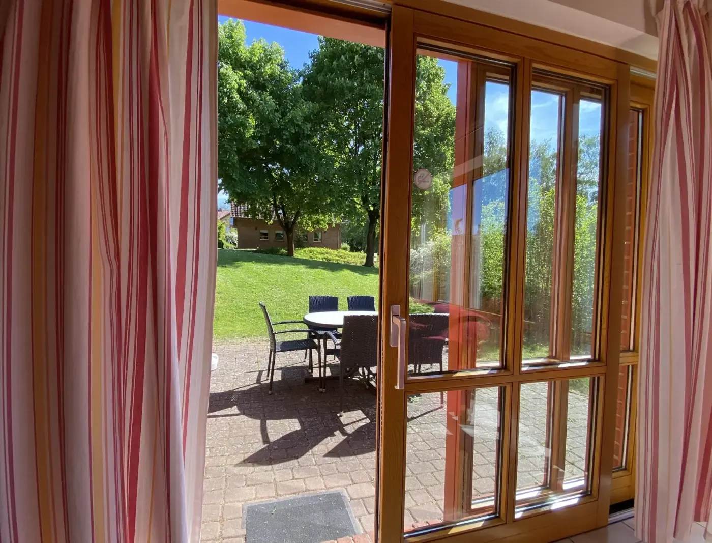 Blick von innen durch eine gläserne Terrassentür auf eine gepflasterte Terrasse mit Essgruppe und einen grünen Garten.