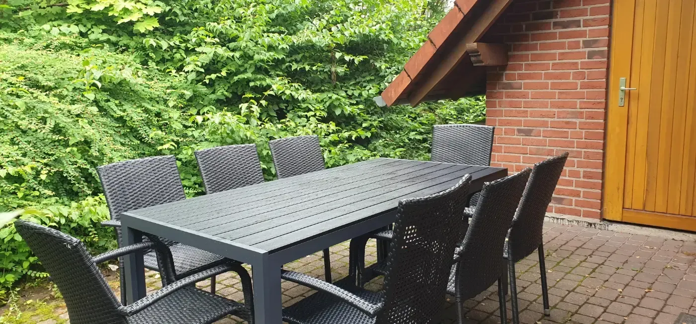 Eine schwarze Essgruppe für den Außenbereich mit einem rechteckigen Tisch und acht geflochtenen Stühlen auf einer Ziegelterrasse neben einem Ziegelhaus.