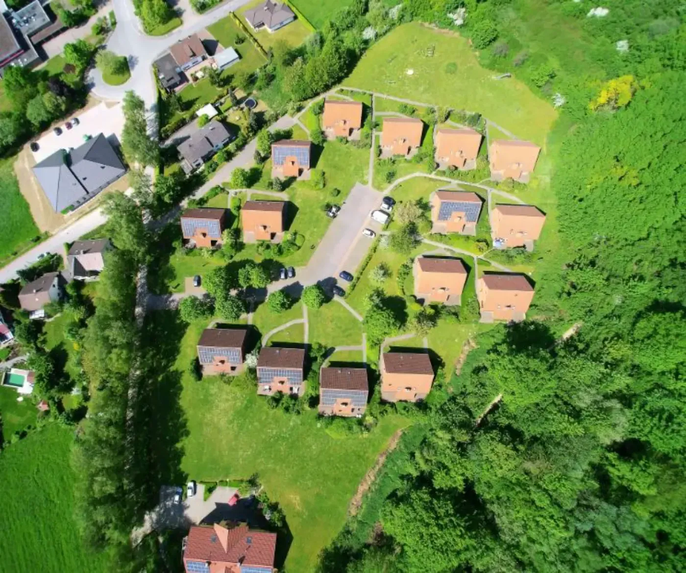 Luftaufnahme einer Wohnsiedlung mit orangebraunen Dächern, umgeben von üppigen grünen Rasenflächen und Wald.