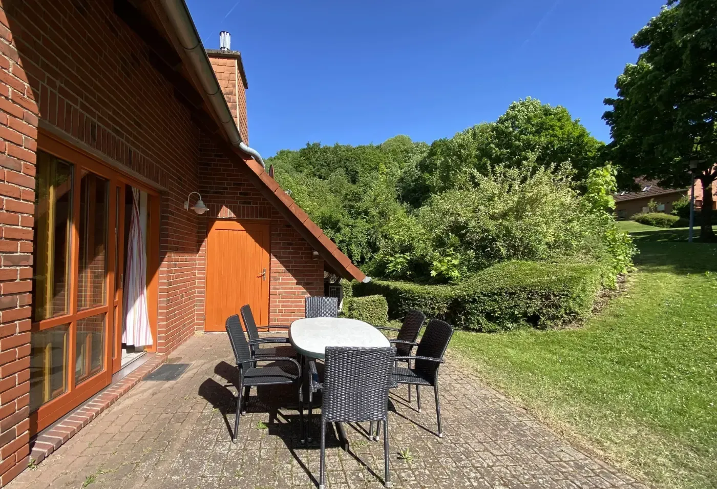 Auf einer Steinterrasse neben einem roten Backsteinhaus mit Blick auf einen grasbewachsenen Hof steht eine Terrassengarnitur mit weißem Tisch und Stühlen.