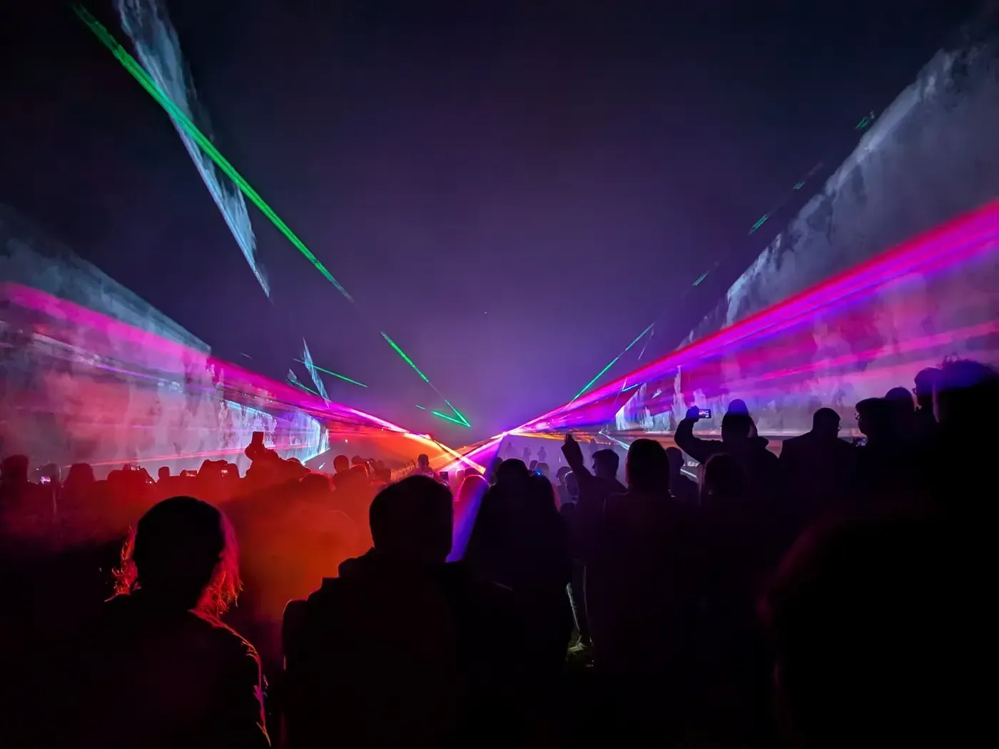 Eine als Silhouette erkennbare Menschenmenge versammelt sich zu einem Konzert, das von leuchtend grünen und pinkfarbenen Laserstrahlen erhellt wird, die den dunklen Himmel durchschneiden.