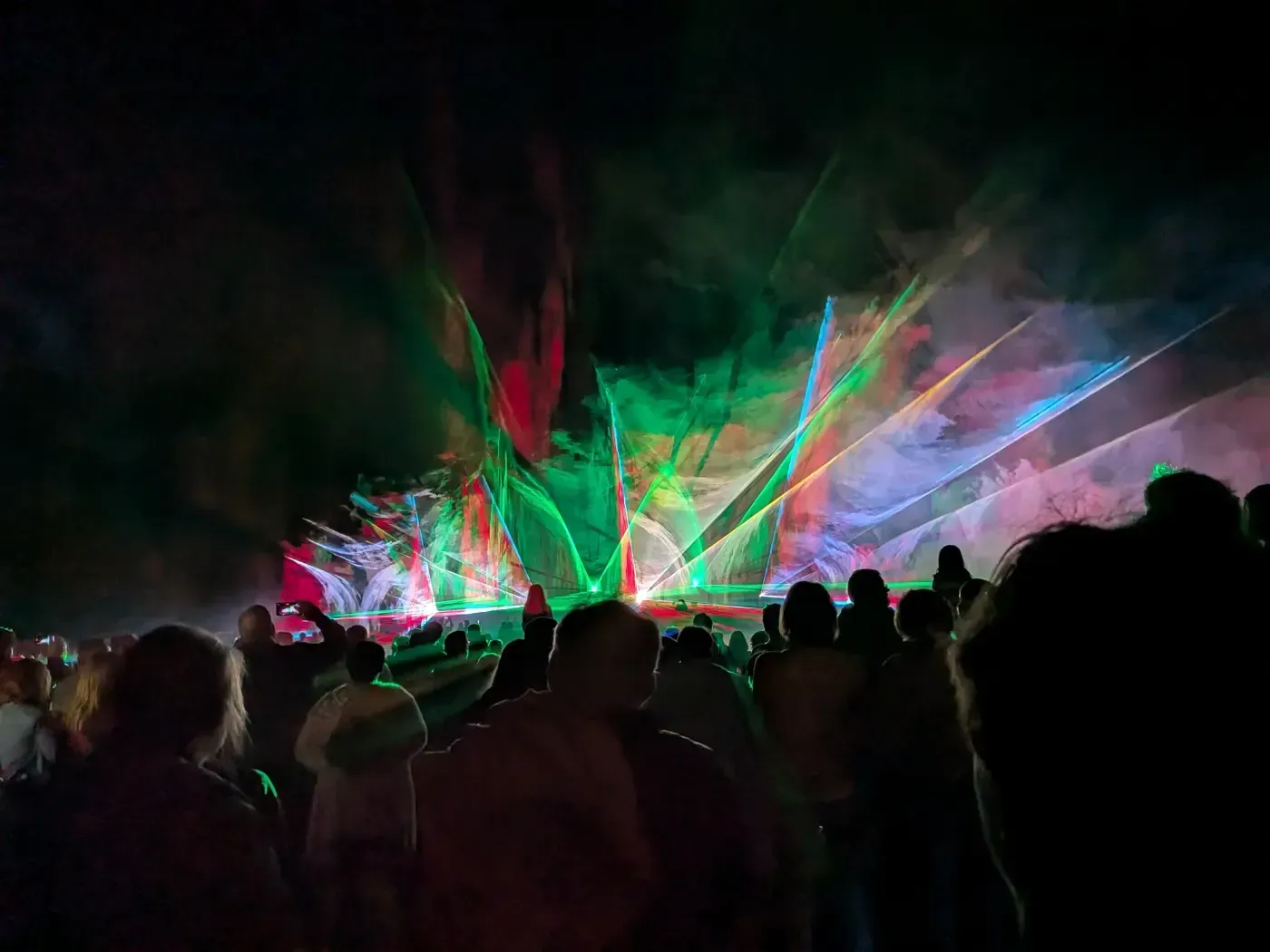 Eine Menschenmenge beobachtet eine nächtliche Lasershow, bei der bunte Laserstrahlen den Rauch vor dem dunklen Himmel erleuchten.