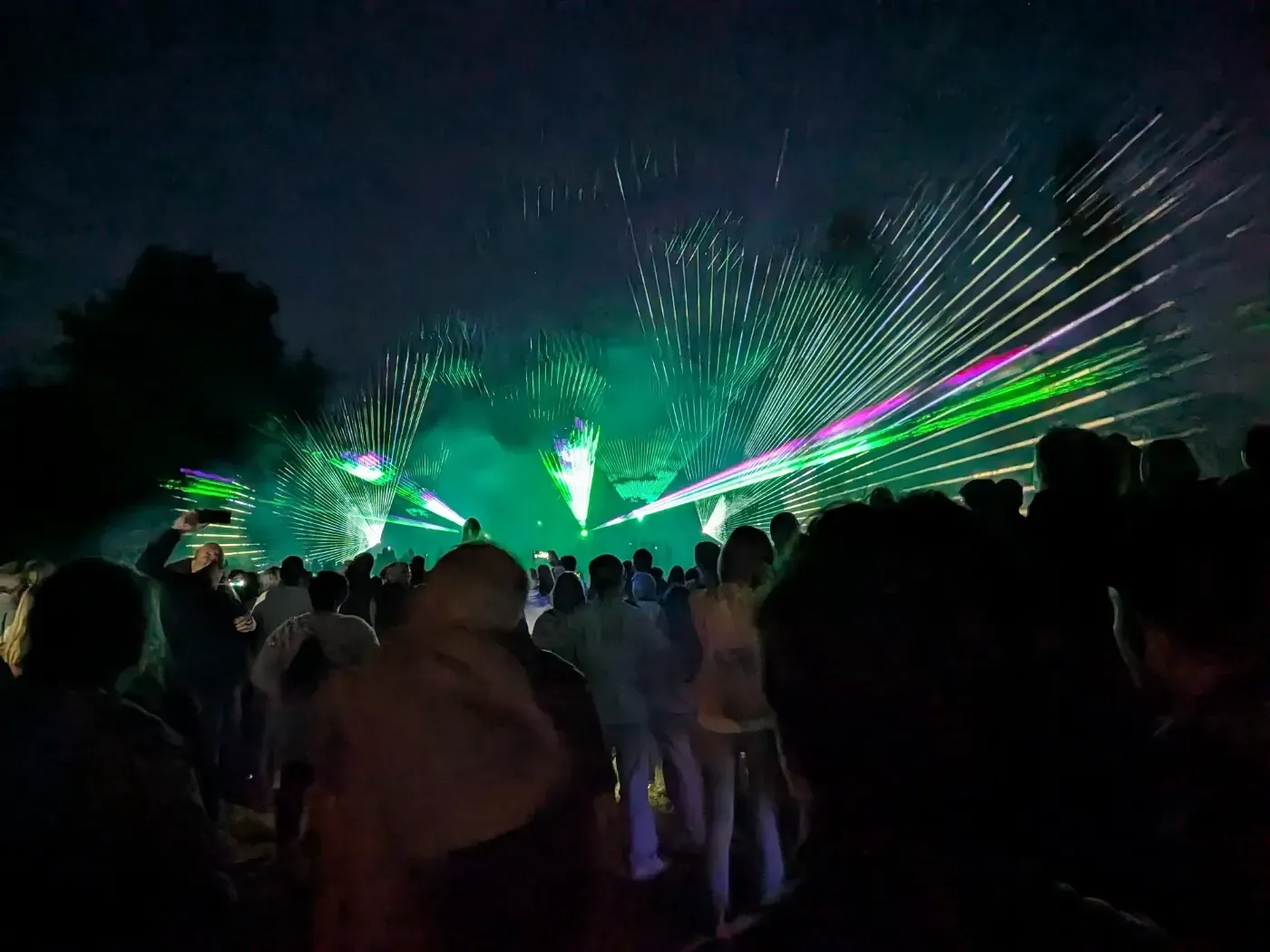 Eine Menschenmenge verfolgt eine farbenprächtige Lasershow am Nachthimmel mit grünen und violetten Lichtstrahlen.