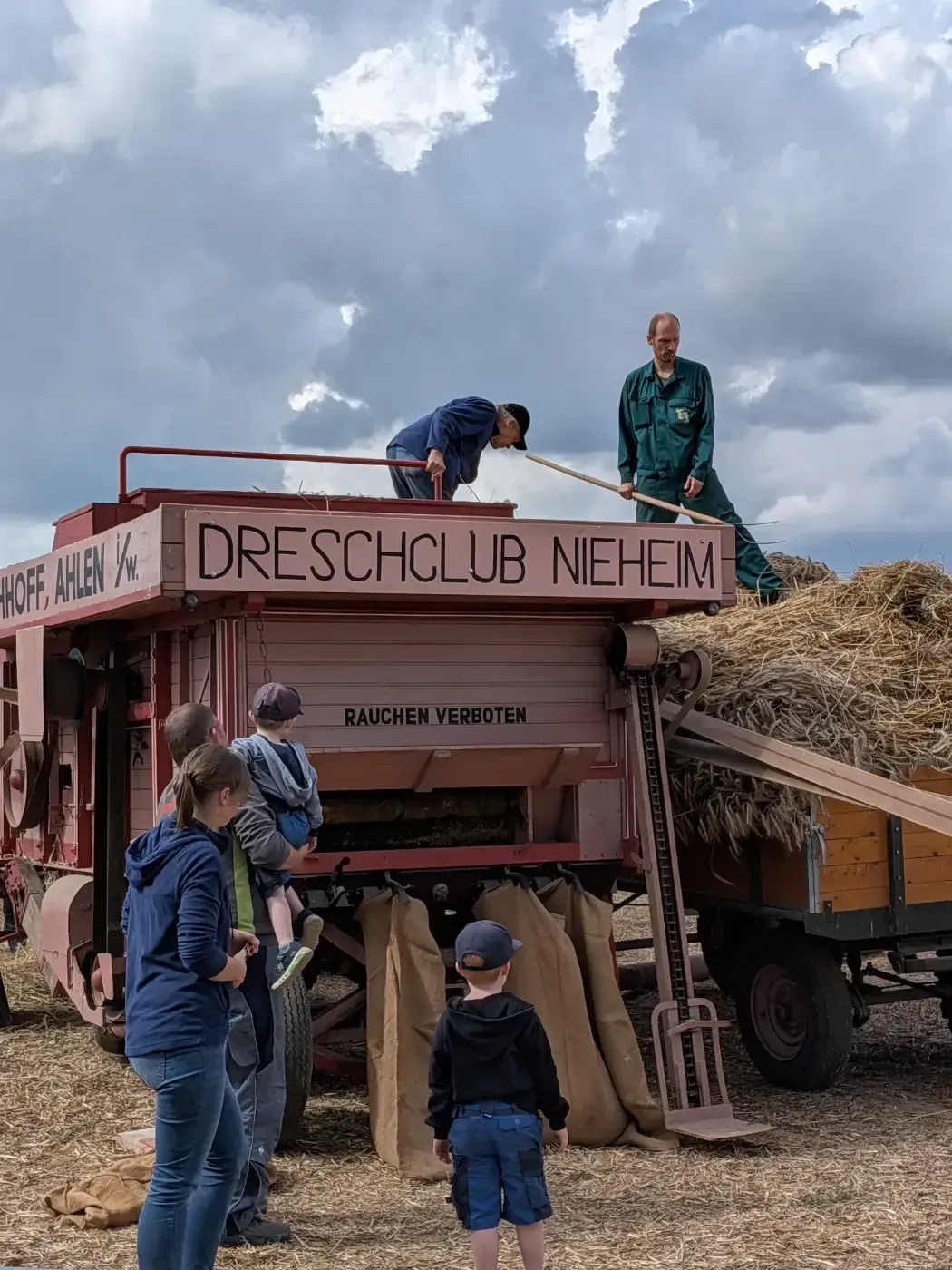 Menschen beobachten eine historische Dreschmaschine mit der Aufschrift „Dreschclub Neheim“ auf einem Feld, während Arbeiter sie mit Stroh befüllen.