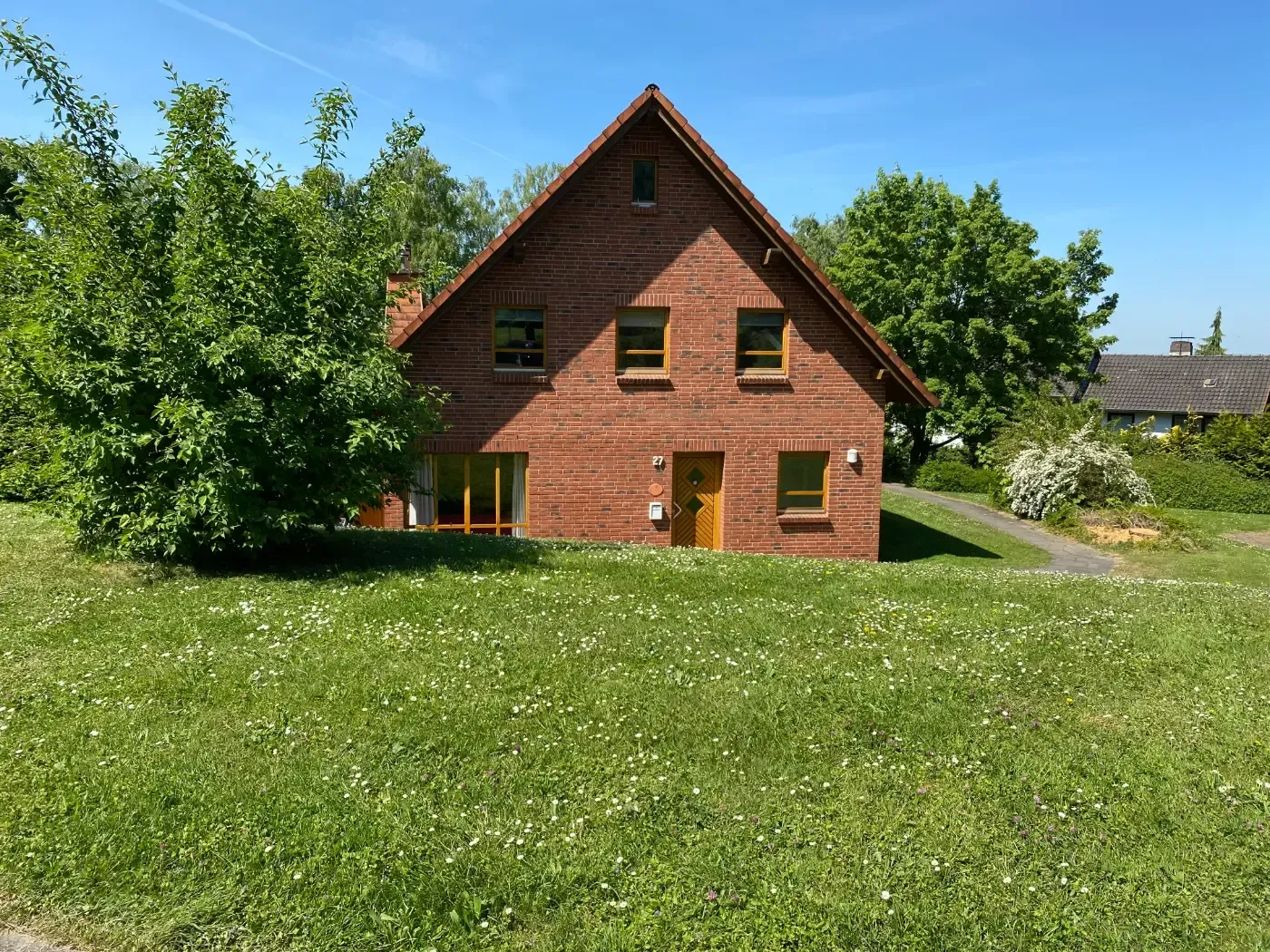Ein zweistöckiges, rotes Backsteinhaus mit Giebeldach, das sich hinter einer großen, sonnenbeschienenen Rasenfläche unter klarem, blauem Himmel befindet.