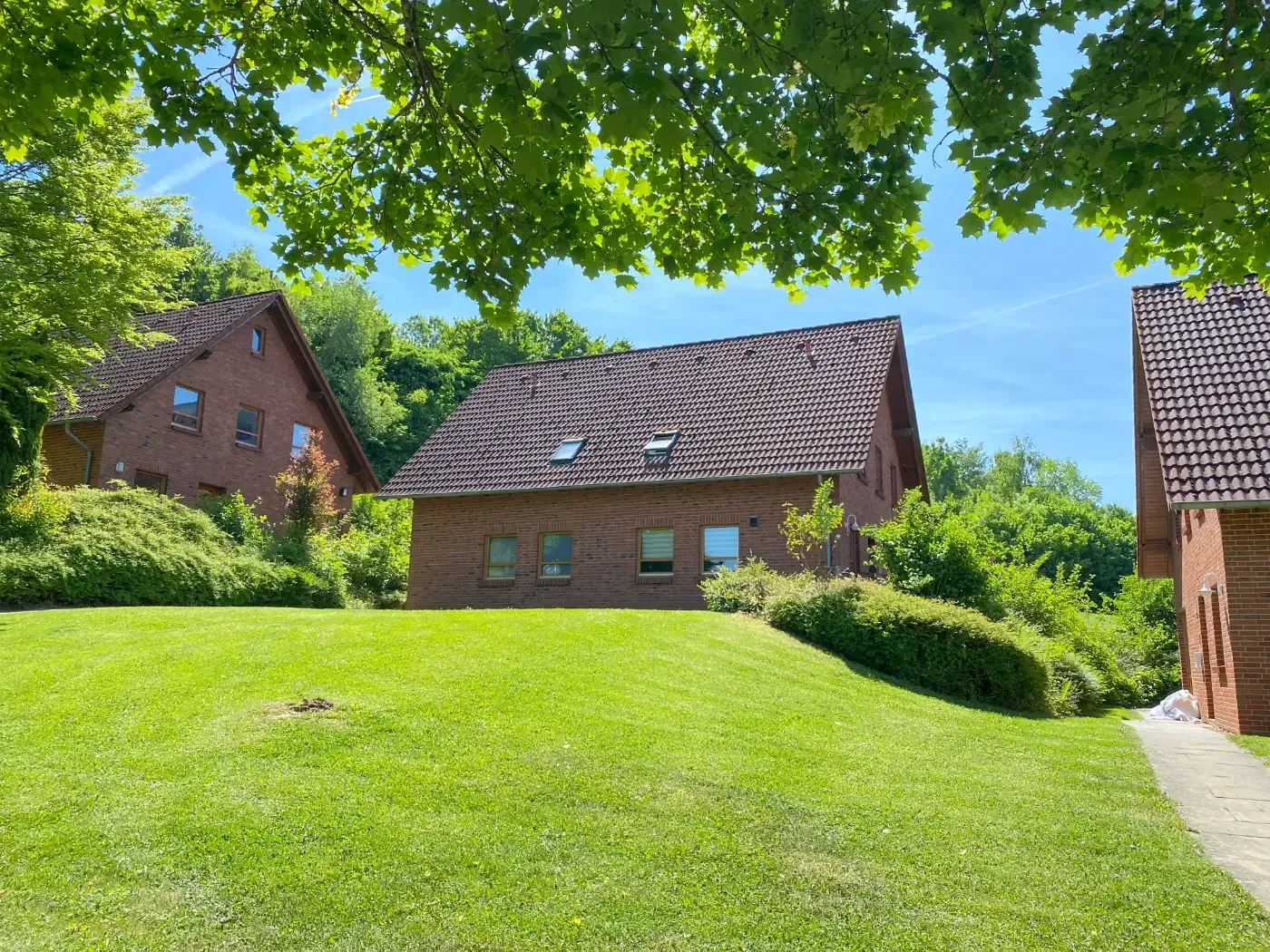 Drei rote Backsteinhäuser mit dunklen Ziegeldächern stehen auf einem grünen, grasbewachsenen Hügel unter strahlend blauem Himmel, eingerahmt von Baumzweigen.
