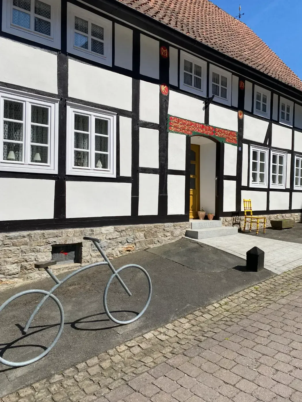 Ein Fachwerkhaus mit weißen Wänden, schwarzen Balken und einer grauen Drahtfahrradskulptur davor an einem sonnigen Tag.