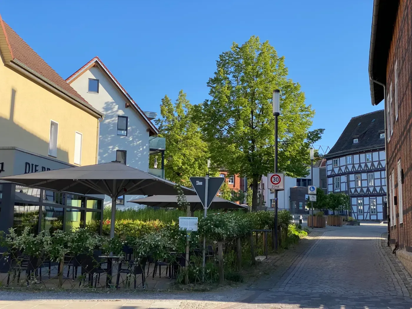Sitzplätze im Freien unter einem großen Sonnenschirm auf einem sonnigen, historischen europäischen Stadtplatz mit Fachwerkhäusern.