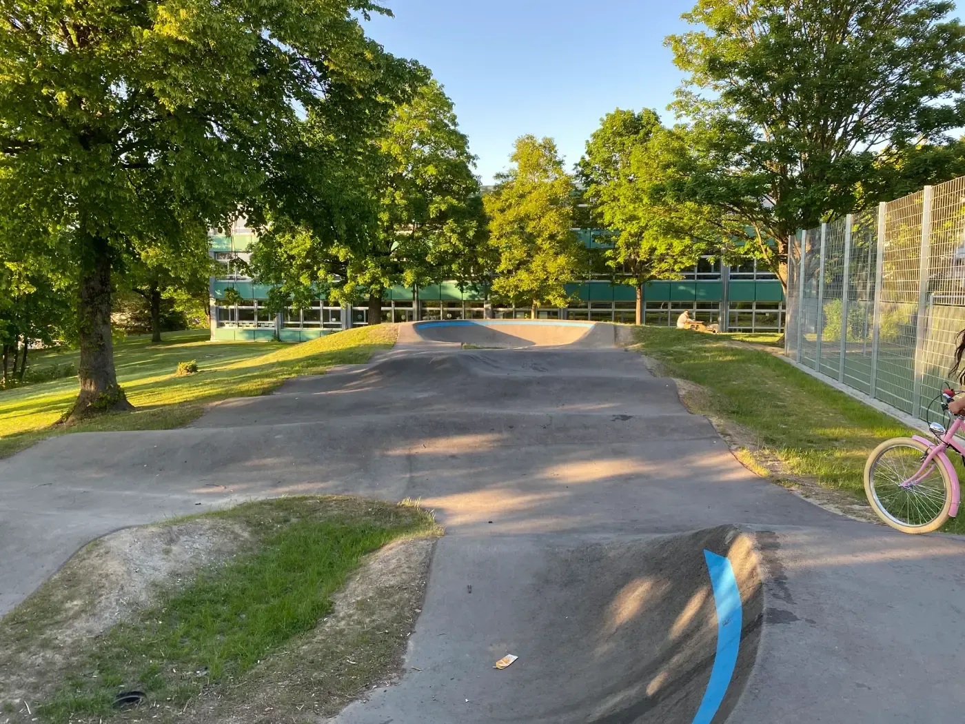 Eine asphaltierte Pumptrack-Anlage für Fahrräder und Roller mit sanften Hügeln und Steilkurven liegt in einem sonnigen Park mit grünen Bäumen.