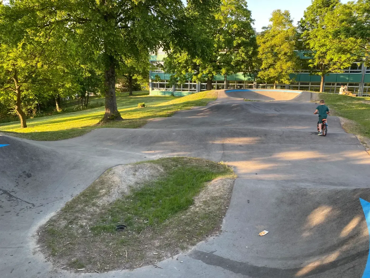 Ein Radfahrer fährt an einem sonnigen Tag auf einer Pumptrack-Anlage aus Beton, die von grünen Bäumen und Gras umgeben ist.