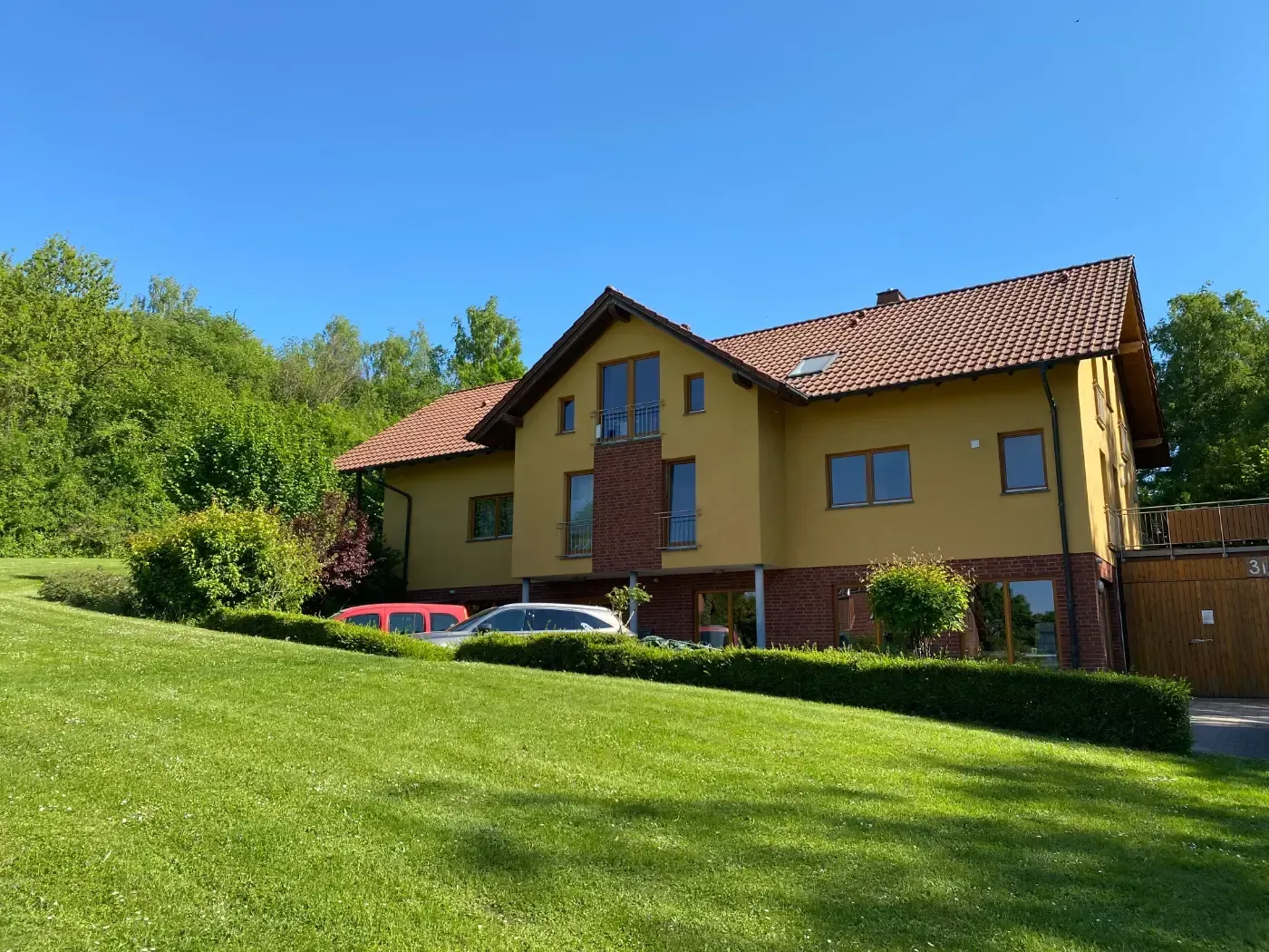 Hinter einer gepflegten Hecke und einem grünen Rasen unter klarem, blauem Himmel steht ein gelbes, zweistöckiges Haus mit braunem Ziegeldach.