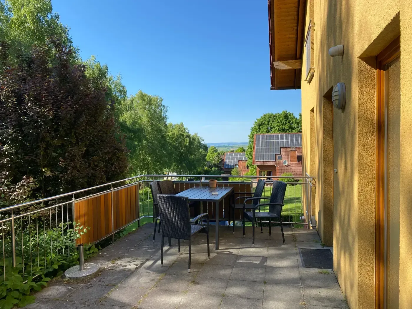 Eine gepflasterte Terrasse mit einem Metalltisch und vier Stühlen sowie einem Metallgeländer mit Blick auf eine sonnige, grüne Landschaft.