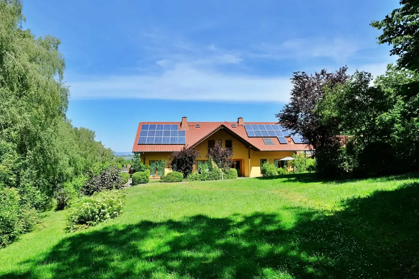 Ein gelbes Haus mit Solarpaneelen auf dem Dach steht auf einer grasbewachsenen Lichtung, umgeben von üppig grünen Bäumen unter blauem Himmel.