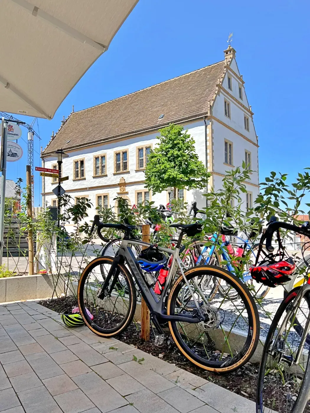 Auf einer Terrasse in der Nähe eines weißen, historischen Gebäudes mit Steindach stehen Fahrräder unter einem klaren blauen Himmel.