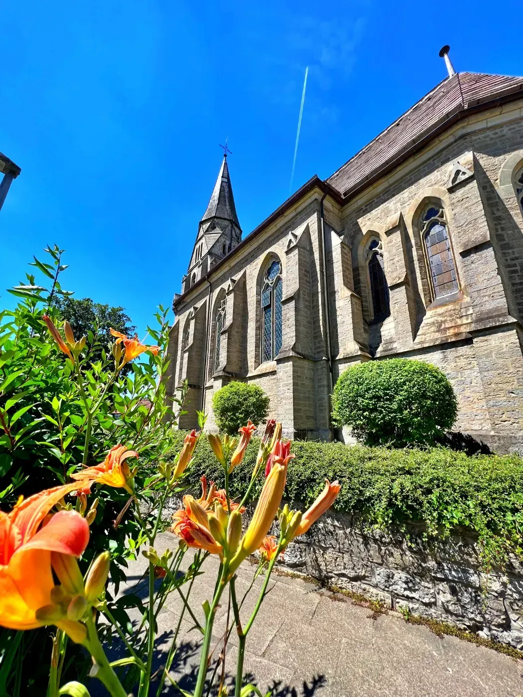 Unter klarem, blauem Himmel steht eine historische Steinkirche mit Kirchturm, im Vordergrund eingerahmt von orangefarbenen Taglilien.